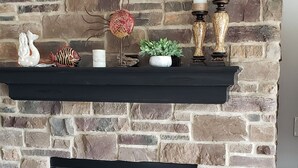Una sala de estar con chimenea de piedra, una repisa negra con adornos, un sofá gris y una mesa de centro con libros.