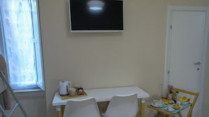 Una habitación pequeña y limpia con un escritorio blanco, dos sillas, un televisor y una mesa de comedor con alimentos para el desayuno.