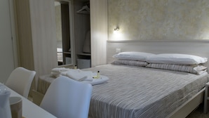 Habitación de hotel con cama, mesita de noche, silla y escritorio. Se observa un perchero con abrigo y una prenda colgada.