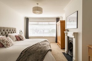 Ein Schlafzimmer mit einer gepolsterten Kopfstütze, einem großen Bett mit gemusterten Kissen, einem Kamin und einem Fenster mit Jalousien.