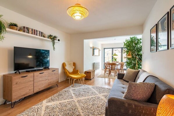 Ein modernes Wohnzimmer mit Flachbildfernseher, Holzschrank, gelbem Sessel, grauer Couch und einem Essbereich mit Tisch und Stühlen.