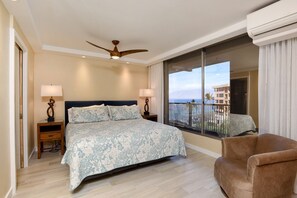 A bedroom with a large bed, two bedside tables with lamps, a ceiling fan, and a view of a balcony with a building and palm trees.