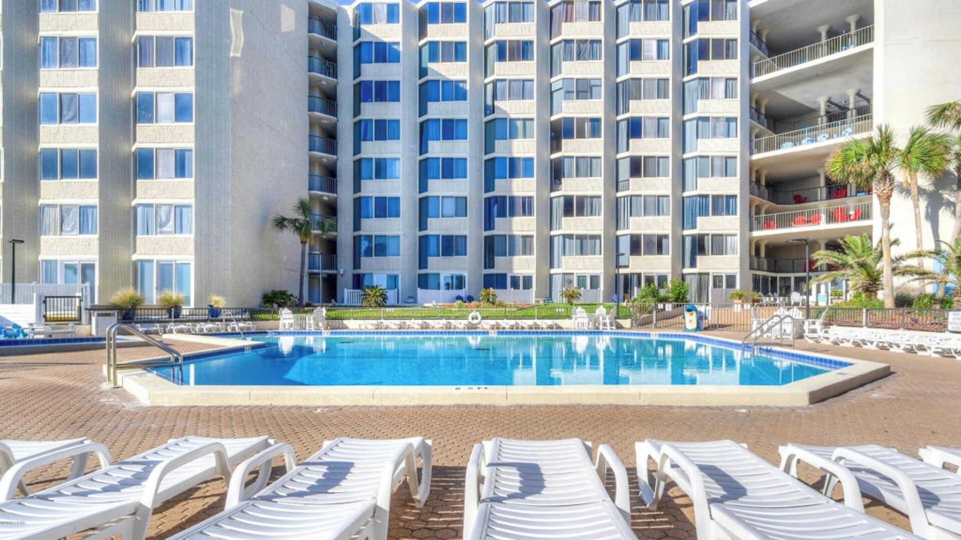 A hotel with a swimming pool, lounge chairs, and a multi-story building with balconies.