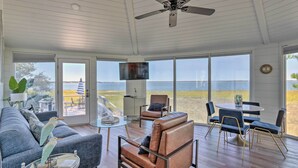 A spacious living room with a large window overlooking a body of water, a ceiling fan, and a television mounted on the wall. - Ocean-View Treetop Escape w/ Fire Pit! (Hilton Head)