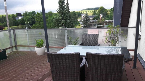 A covered outdoor area with a table and chairs, surrounded by a glass railing, overlooking a scenic landscape with trees and buildings. - Vacation apartment, good location in the mining region Erzgebirge, mountain town Schneeberg (Schneeberg, Sachsen, Deutschland)