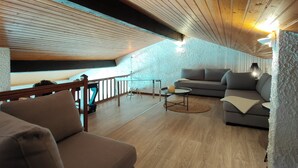 A cozy loft bedroom with a sloped ceiling, wooden beams, a grey sofa, a small round table, and a glass railing overlooking a balcony. - Air-conditioned "Carmen" villa with heated pool near the ocean (Biscarrosse)