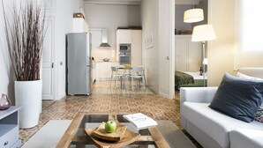 Un salon moderne avec une table basse en verre, un canapé blanc et un coin repas avec une cuisine en arrière-plan.