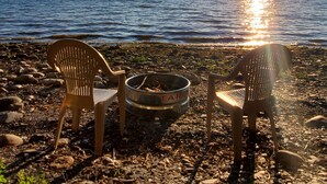 Two chairs and a metal container on a rocky beach with the sun setting over the water. - Lake Champlain Waterfront: Amazing Sunsets, Private Beach, and Firepit (Swanton)