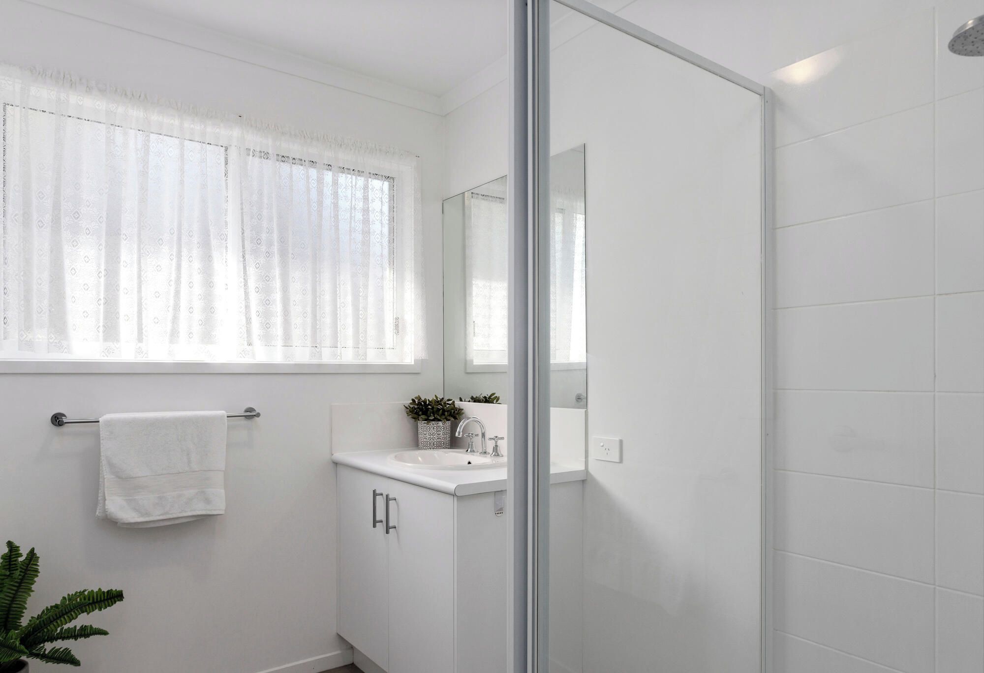 A bathroom with a glass shower door, a white sink, and a window with patterned curtains.