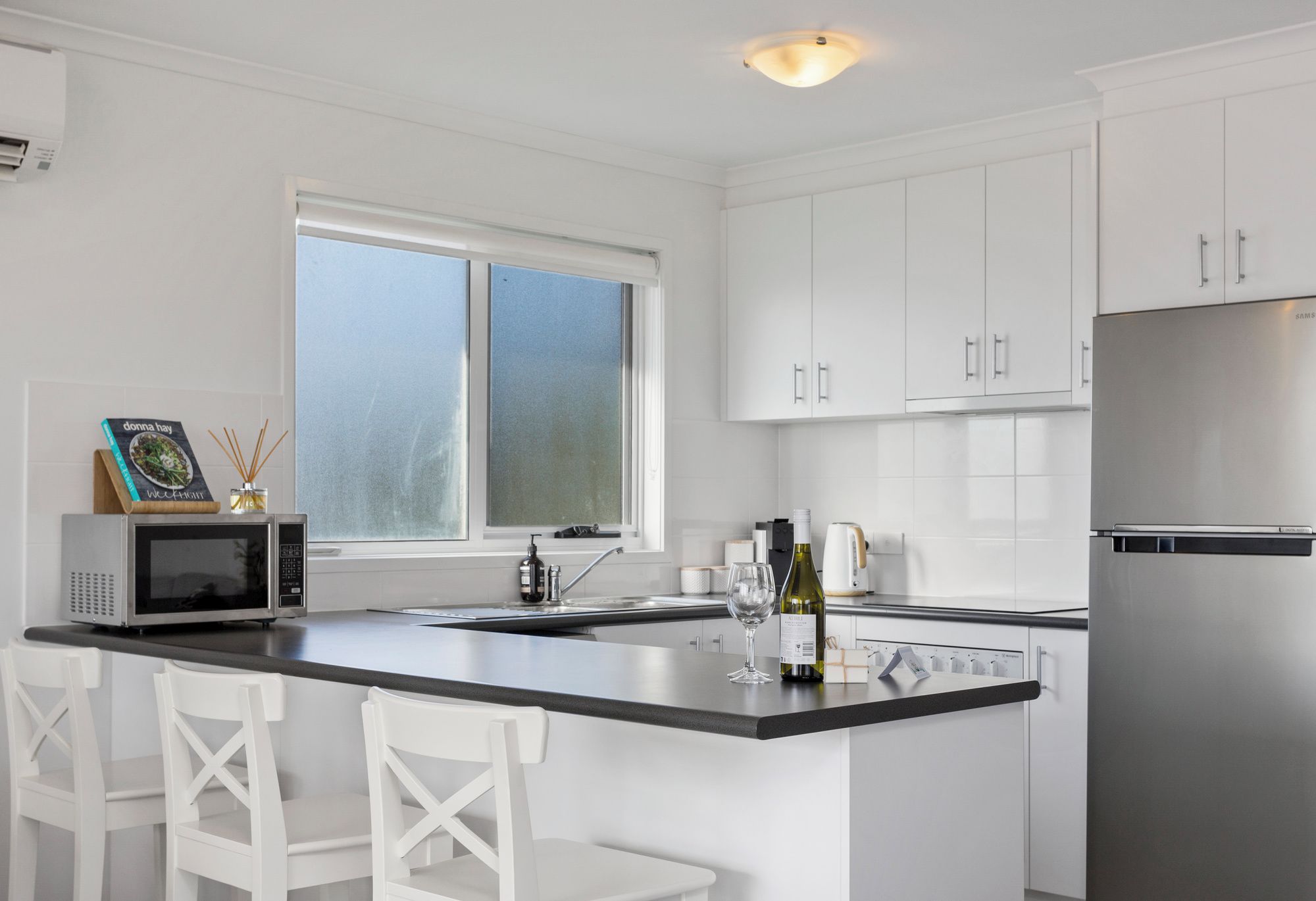 A modern kitchen with white cabinetry, a stainless steel refrigerator, a microwave, and a large window.