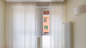 A room with a view through sheer curtains to an outdoor area with a radiator and a window with shutters. - San Felice 135 - 3 (Bologna)