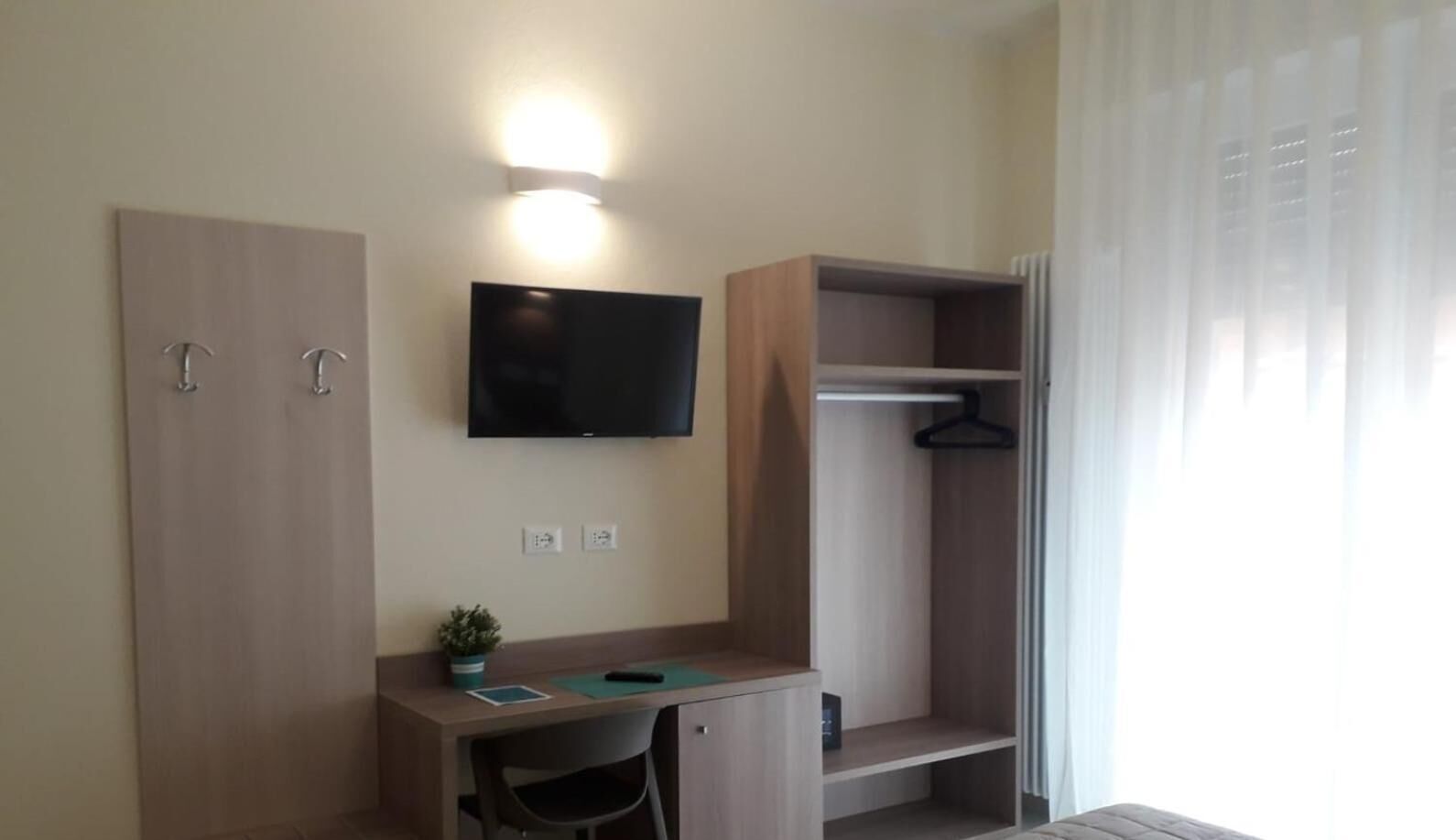 A compact room with a desk, a TV mounted on the wall, a coat closet, and a window with curtains.