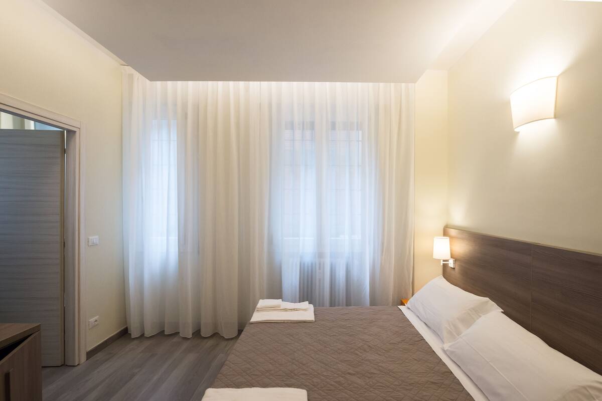 Una camera d'albergo con un letto, tende, una lampada a parete e una porta.
