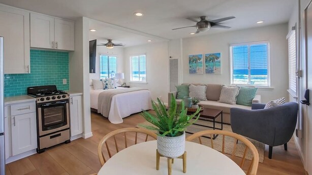 A modern kitchen with white cabinets, a stove, and a green backsplash. There is a dining area with a table and chairs, a sofa, and a ceiling fan.