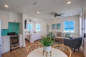A modern kitchen with white cabinets, a stove, and a green backsplash. There is a dining area with a table and chairs, a sofa, and a ceiling fan. - Pacific House #1 - Extreme Oceanfront (Oceanside)