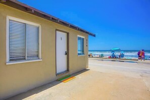 A building with a door and windows, a beach in the background, and people and beach chairs.