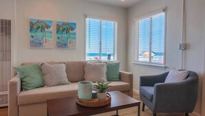 A living room with a beige sofa, a dark armchair, a coffee table with a plant, and two beach-themed paintings on the wall.
