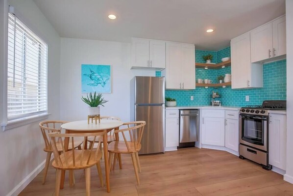 A kitchen with white cabinets, a stainless steel refrigerator, a built-in oven, and a dining area with wooden chairs. - Pacific House #1 - Extreme Oceanfront (Oceanside)