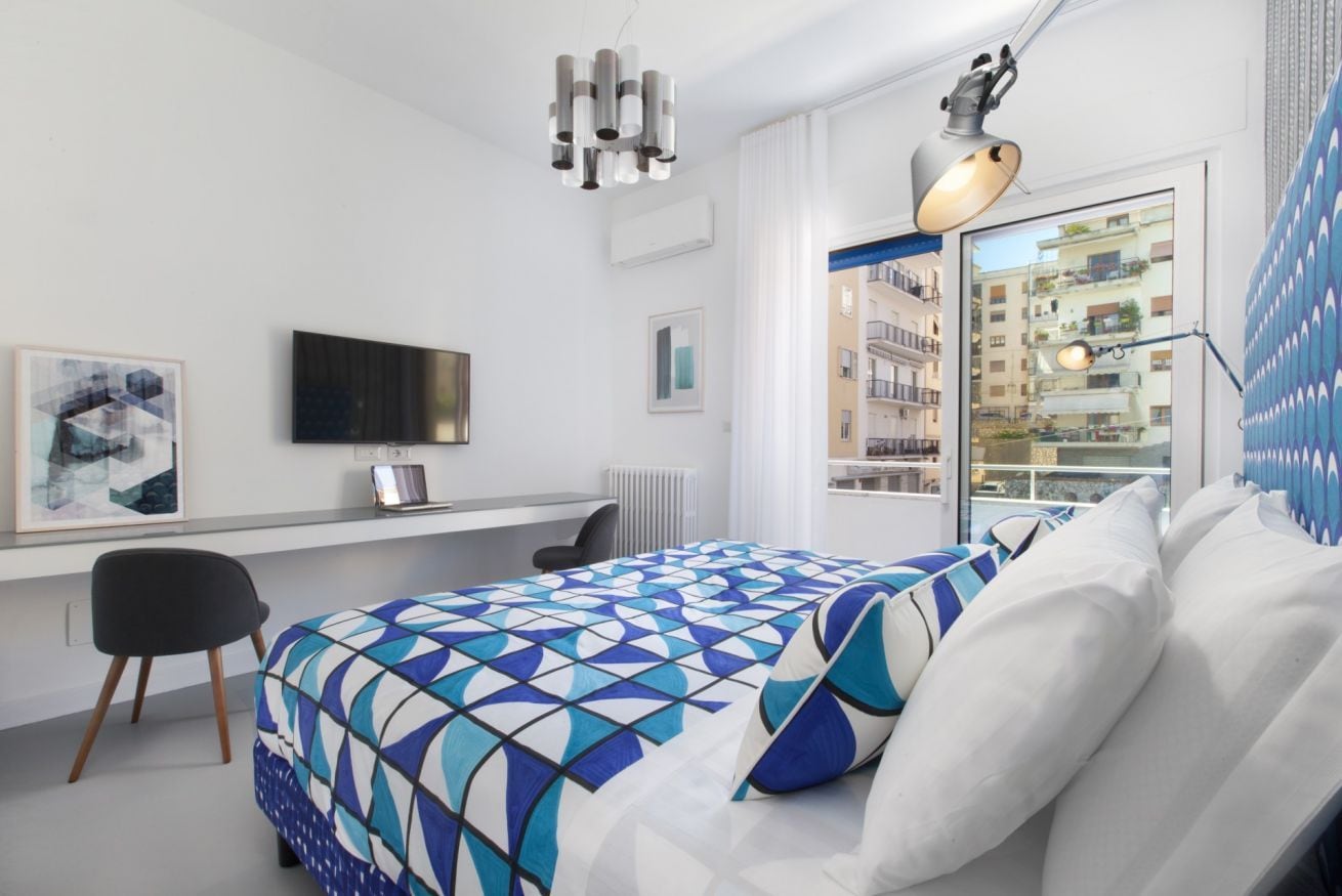 A modern bedroom with a geometric-patterned bedspread, a desk with a laptop, a television, and a view of a residential building through a window.