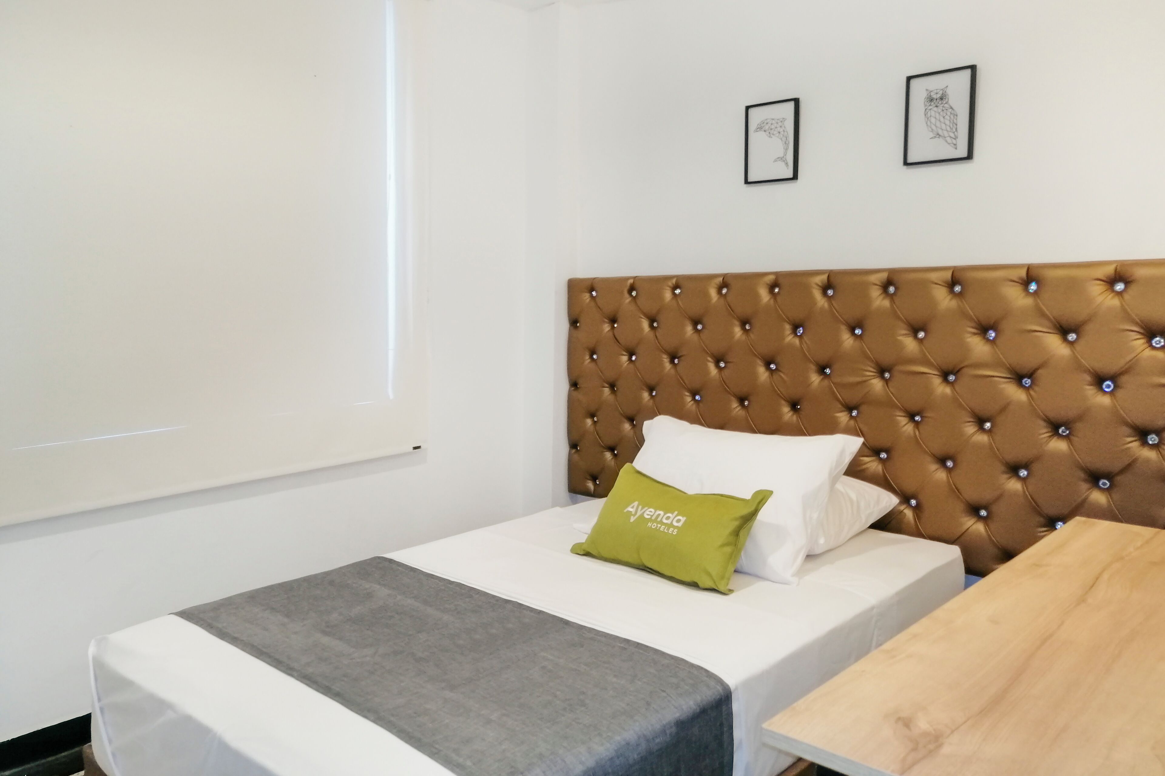 Habitación de hotel con cabecera capitonada, cama con ropa blanca y almohada verde, una mesita y dos cuadros en la pared.