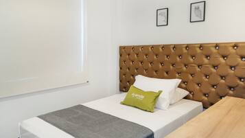 Habitación de hotel con cabecera capitonada, cama con ropa blanca y almohada verde, una mesita y dos cuadros en la pared.