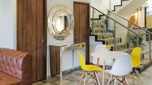 Een eethoek met een witte tafel, geel-witte stoelen, een bruine leren bank, een houten deur en een trap met een glazen balustrade.