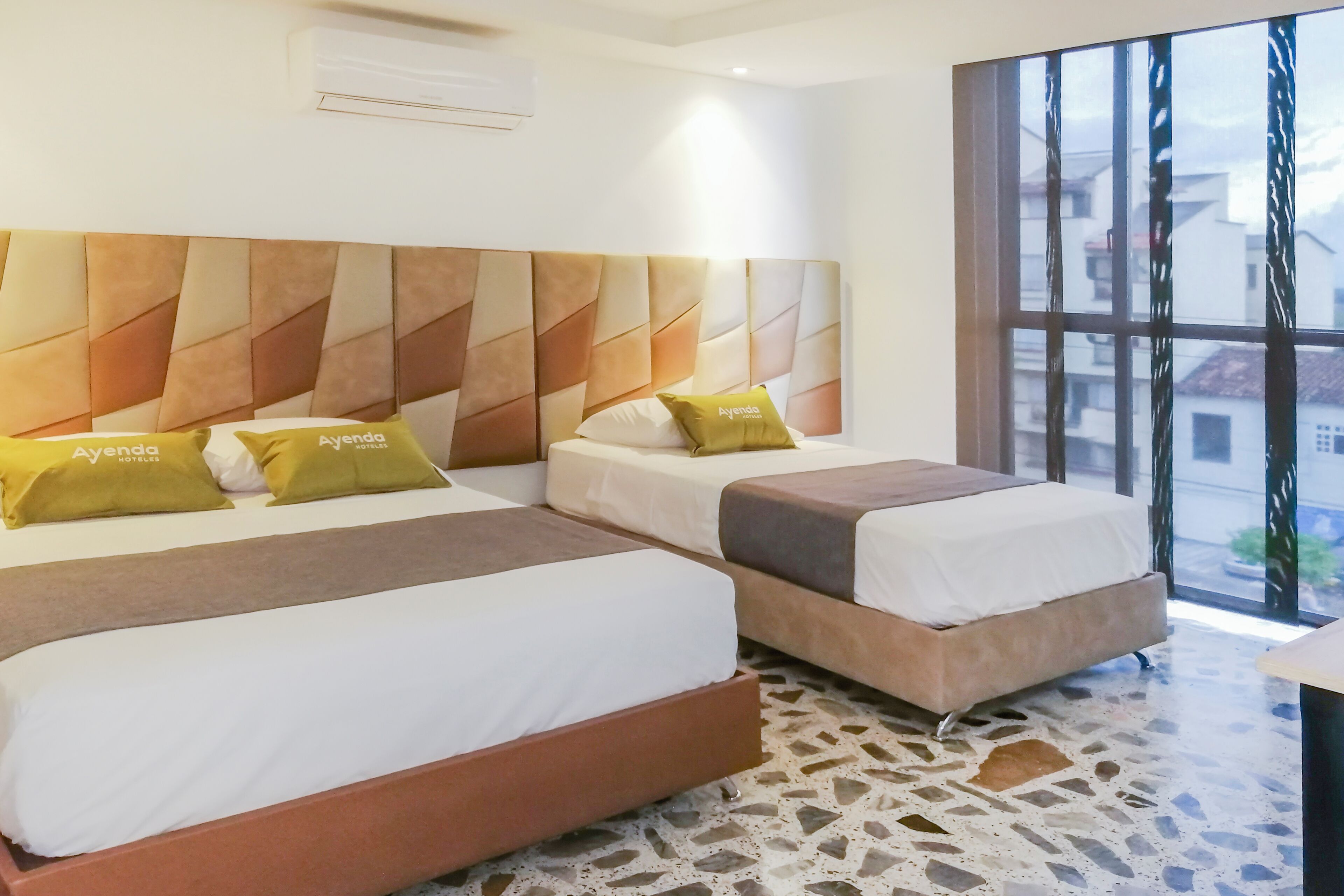 Une chambre d’hôtel avec deux lits, une tête de lit à motifs et une grande fenêtre donnant sur des immeubles.
