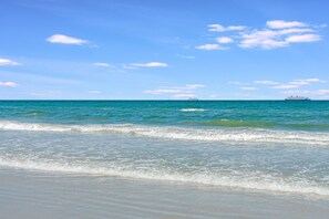 Een strand met helderblauw water en een schip in de verte.