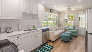 A modern kitchen with white cabinets, a dishwasher, and a toaster. There is a beige sofa and a teal ottoman in the living area. - Sizzling Prices-COOL Summer Retreat (Cocoa Beach)