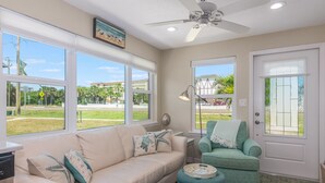 A living room with a beige sofa, teal ottoman, and a green armchair. There are large windows, a ceiling fan, and a door with a glass panel. - Sizzling Prices-COOL Summer Retreat (Cocoa Beach)