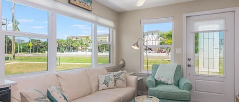 A living room with a beige sofa, teal ottoman, and a green armchair. There are large windows, a ceiling fan, and a door with a glass panel.