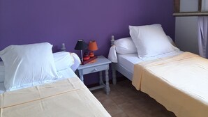 Huoneessa on kaksi sänkyä, violetit seinät, pieni pöytä, jossa on lamppu, ja ikkuna, jossa on verhot.
