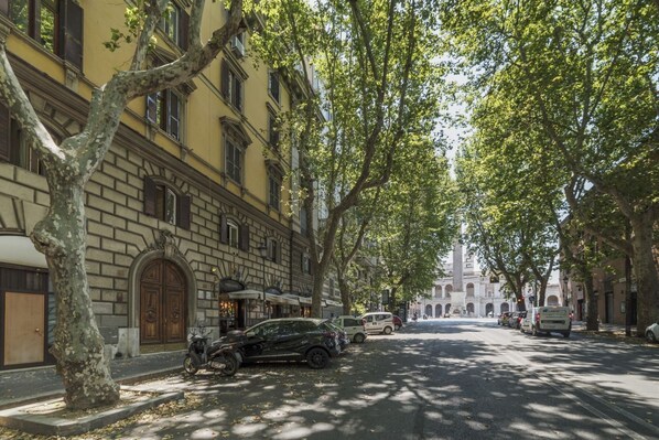 A tree-lined street with parked cars and a building with a large arched entrance.