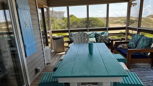 Une terrasse extérieure couverte, avec une table turquoise et des chaises blanches, un canapé agrémenté de coussins à motifs, et une vue sur la plage et les bâtiments en arrière-plan.