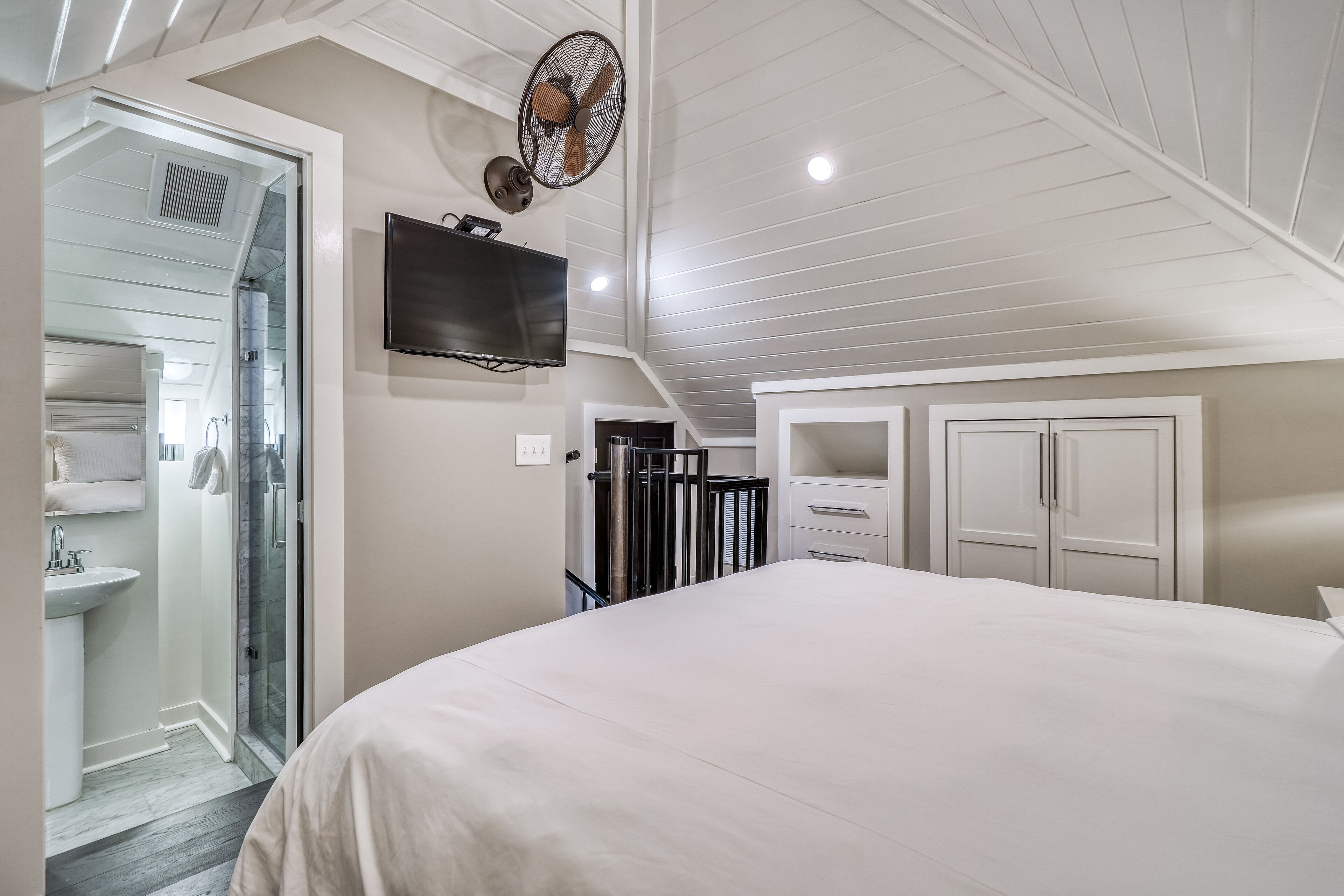 A bedroom with a large bed, a TV mounted on the wall, and a ceiling fan.