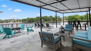 A covered outdoor seating area with chairs and tables, overlooking a body of water and greenery. - Luxury A-Frame on Lake LBJ, Wakesurf Paradise, Two-Story Dock, Sleeps 16 (Kingsland)