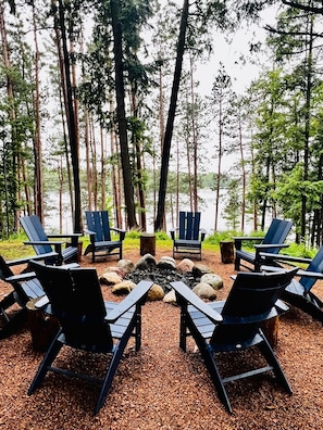 A forested area with a fire pit surrounded by Adirondack chairs facing a lake. - Little Fork Lodge - Three Lakes Lakefront on the trails with Firepit/Game Room (Three Lakes)