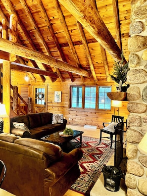 A cozy cabin interior with a wooden ceiling, a stone wall, a fireplace, a couch, a coffee table, and a rug.