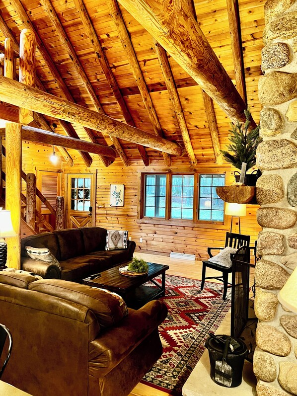 A cozy cabin interior with a wooden ceiling, a stone wall, a fireplace, a couch, a coffee table, and a rug.