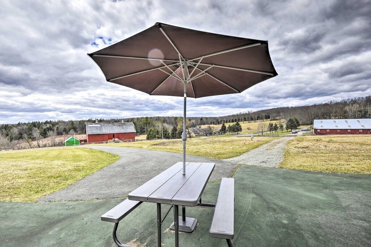 An outdoor picnic area with a table and bench, a large umbrella, and a rural landscape in the background.