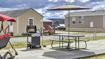 A picnic setup with a grill, table, and umbrella in a residential area with houses and a car in the background.