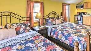 A bedroom with two beds, a nightstand, a lamp, a window with curtains, and a kitchenette with a microwave and stove.