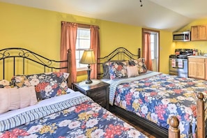 A bedroom with two beds, a nightstand, a lamp, a window with curtains, and a kitchenette with a microwave and stove. - Double Retreat · Poconos Nature Escape (Pleasant Mount)
