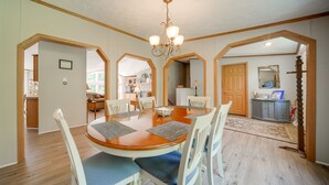 Un comedor con mesa de madera y sillas blancas, una araña y vista a la cocina.