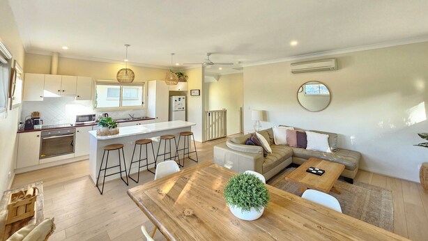 A modern kitchen with white cabinets, a central island with stools, and a dining area with a wooden table and chairs. - Summer House - Cosy, comfortable, located close to town (Robe)