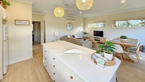 A modern kitchen with a central island, a dining area with a wooden table and chairs, a TV, and a large window providing natural light. - Summer House - Cosy, comfortable, located close to town (Robe)