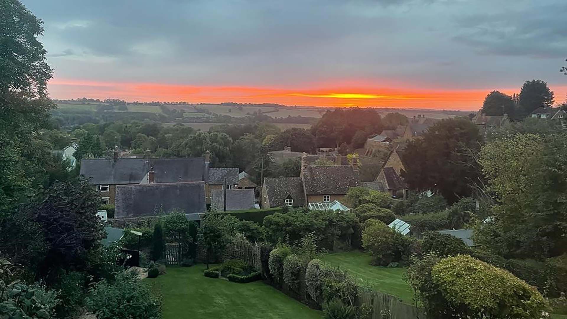 A rural village with traditional stone houses, surrounded by lush greenery and a sunset in the background.