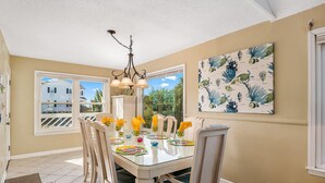 A dining area with a glass table, chairs, and a chandelier. There is a window with a view of houses and trees, and a wall with a botanical print. - Second Row, 4 Bedroom/3 Bath Home w/ Private Pool and Dock. Sleeps 10 (Holden Beach)