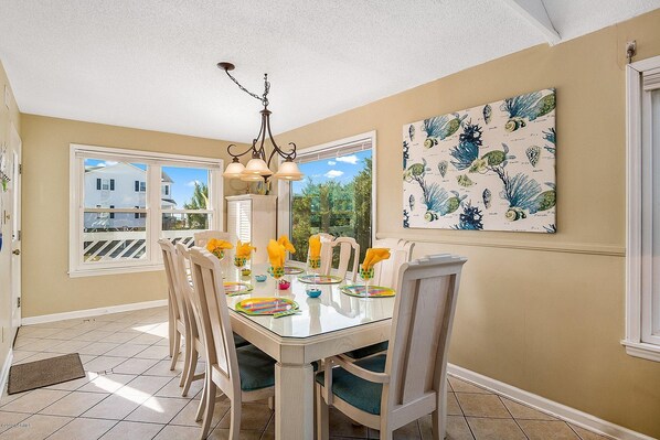 A dining area with a glass table, chairs, and a chandelier. There is a window with a view of houses and trees, and a wall with a botanical print.