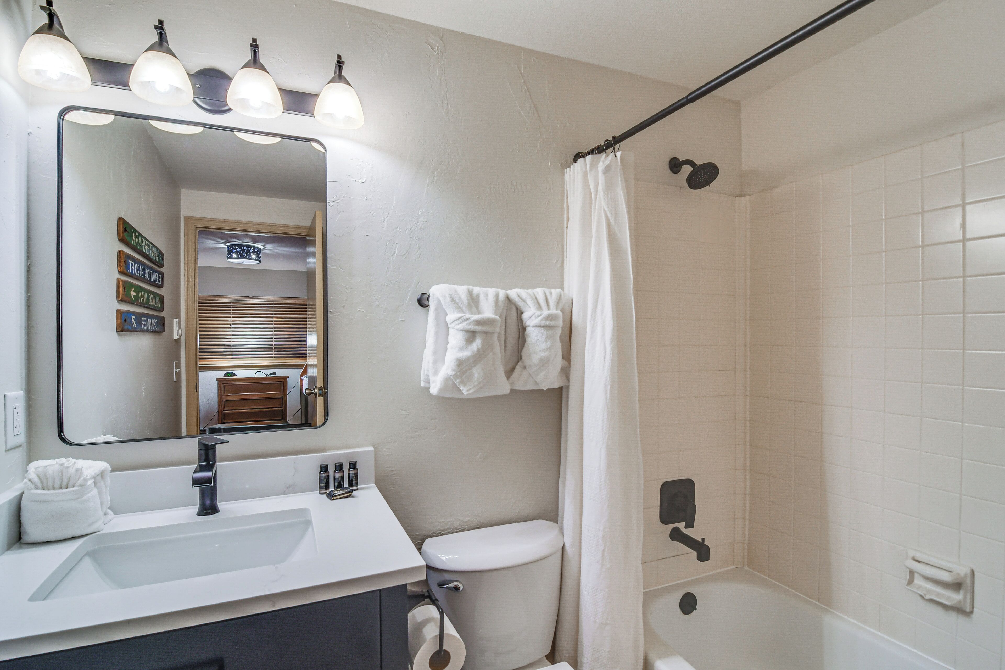 Un baño con lavamanos blanco, espejo e inodoro. Hay toallas colgadas en la pared y una cortina de ducha.